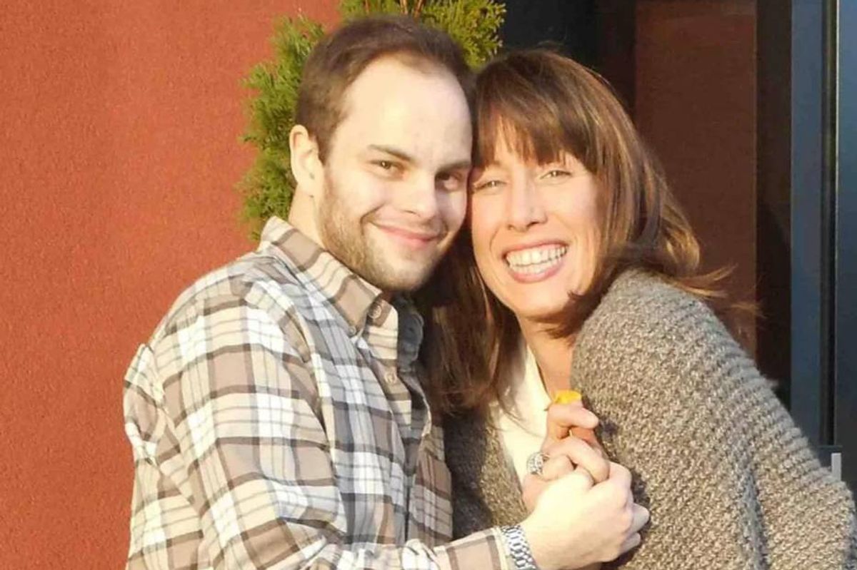 Two individuals are standing close together, smiling, and embracing each other. The individual on the left is wearing a plaid shirt, while the individual on the right is dressed in a light-colored jacket. The background is a solid red wall.