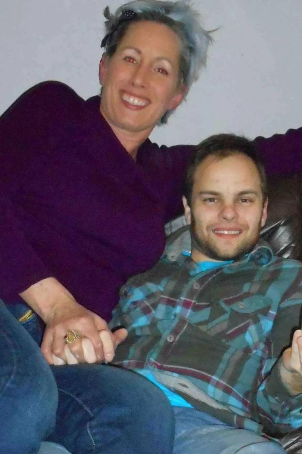 An individual with a beard and mustache is sitting on a couch, wearing a blue plaid shirt, while another person in a purple top stands behind them, extending an arm in a gesture of support or affection. The setting appears to be an indoor environment, possibly a living room.