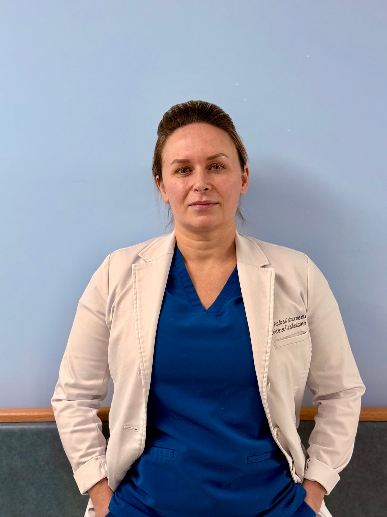 A physician in a white lab coat and blue shirt stands next to a blue wall.