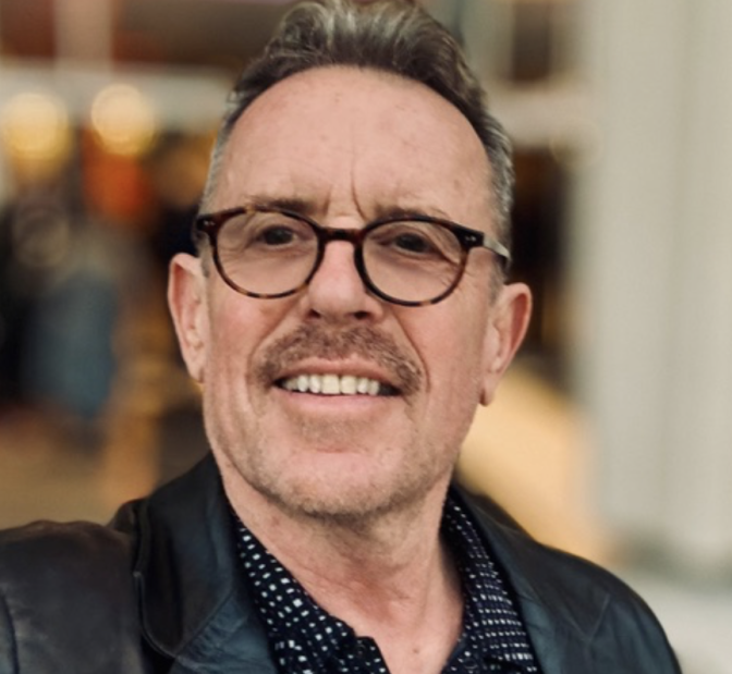 A light skinned man with greying hair, wearing tortoiseshell glasses, smiles at the camera.