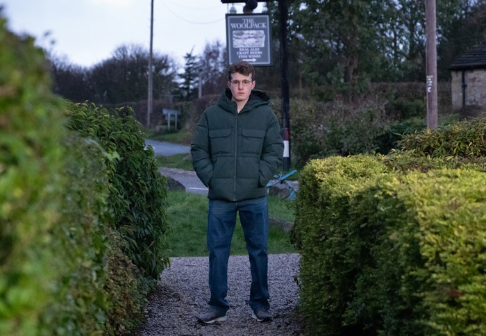 Arthur stands alone at the gate of Mulberry Cottage, under the sign of The Woolpack, in a scene from Emmerdale