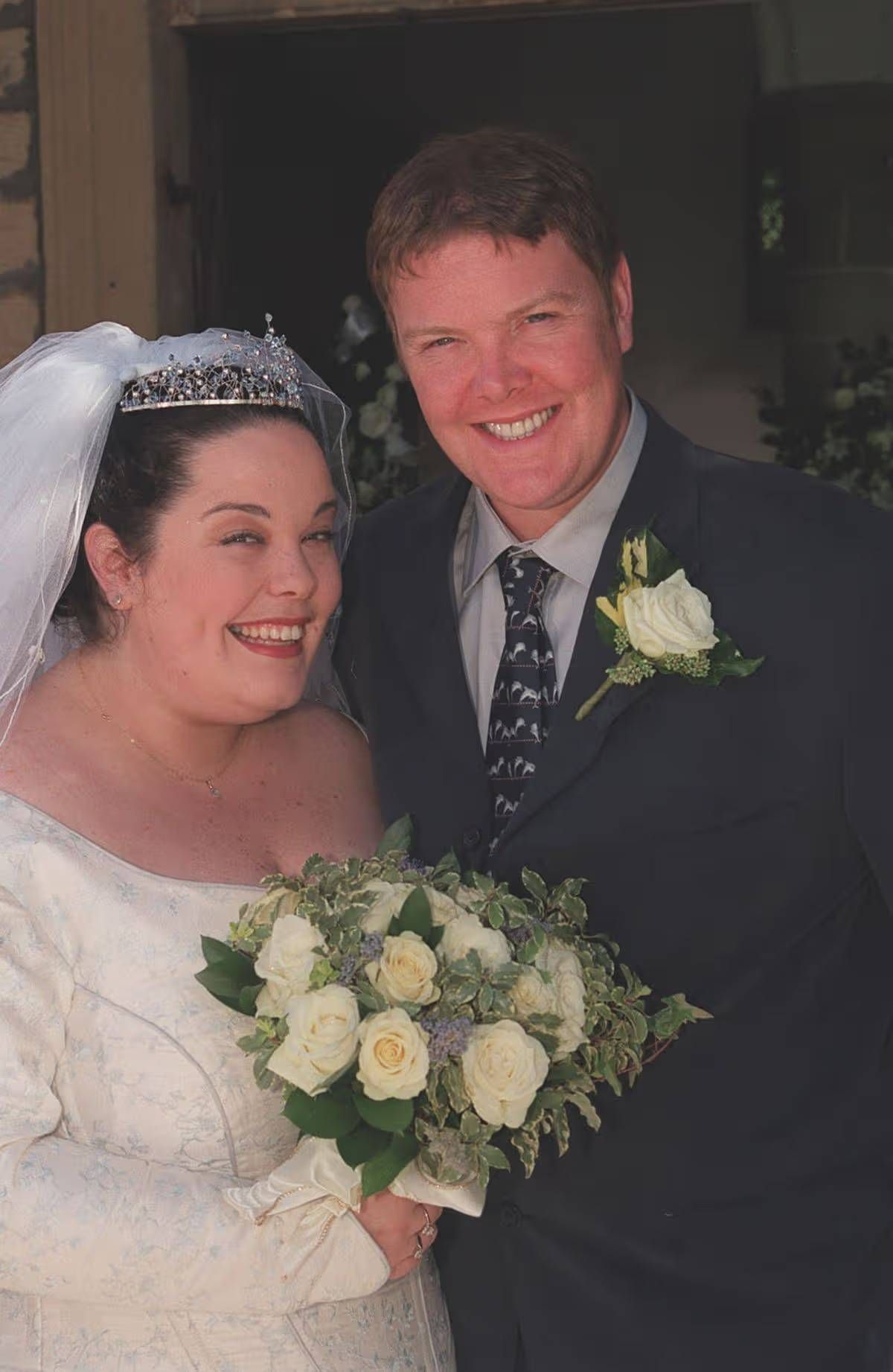 An image depicting a newlywed couple standing close together, likely at their wedding. The woman, dressed in a wedding gown and veil, is beaming with joy, while the man, wearing a suit and tie with a boutonnière, shares a warm and affectionate smile. The background is blurred, focusing the attention on the couple's expressions and their joyous union.