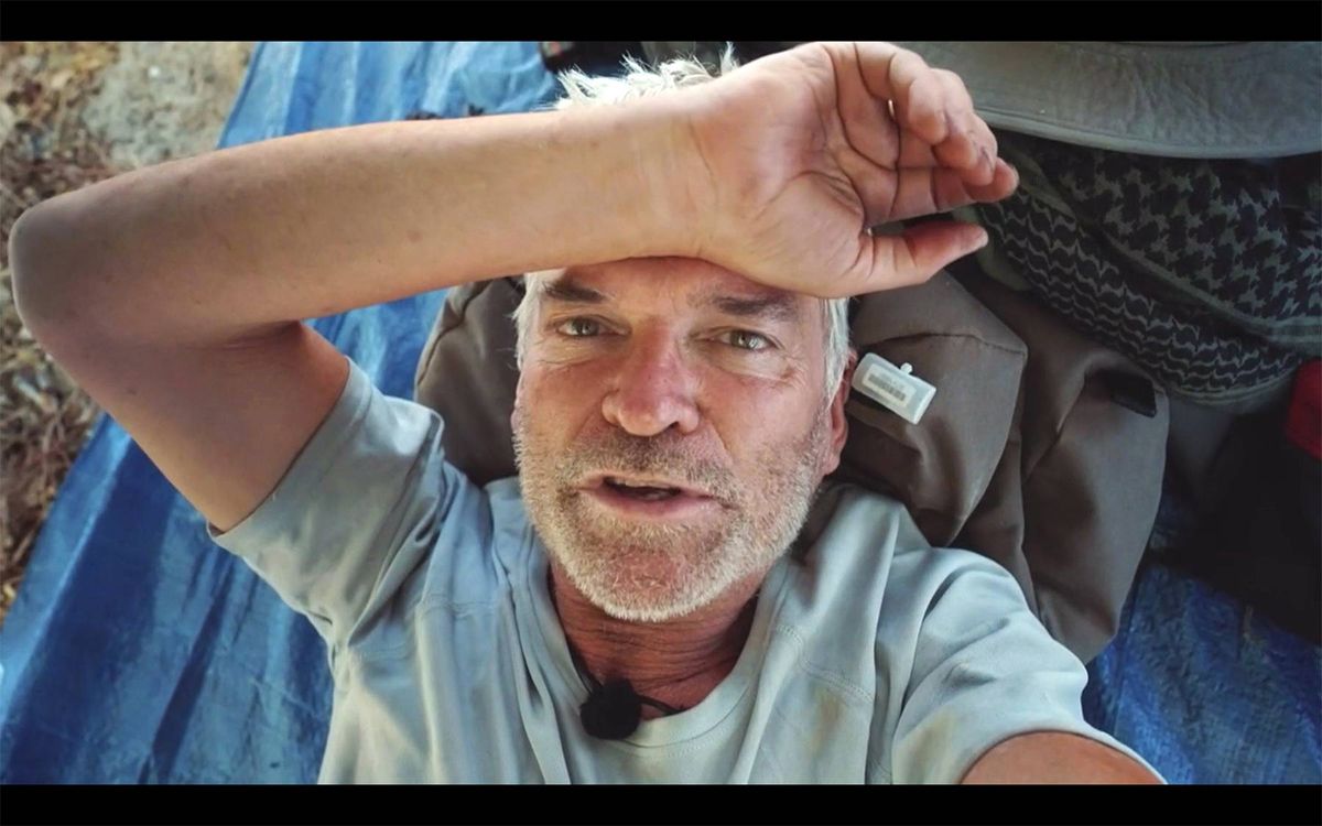 An adult male is seen with one hand resting on his forehead, appearing to be in a moment of contemplation or discomfort. He is dressed in a light-colored t-shirt, and the background is intentionally blurred, suggesting a focus on his expression and gesture.