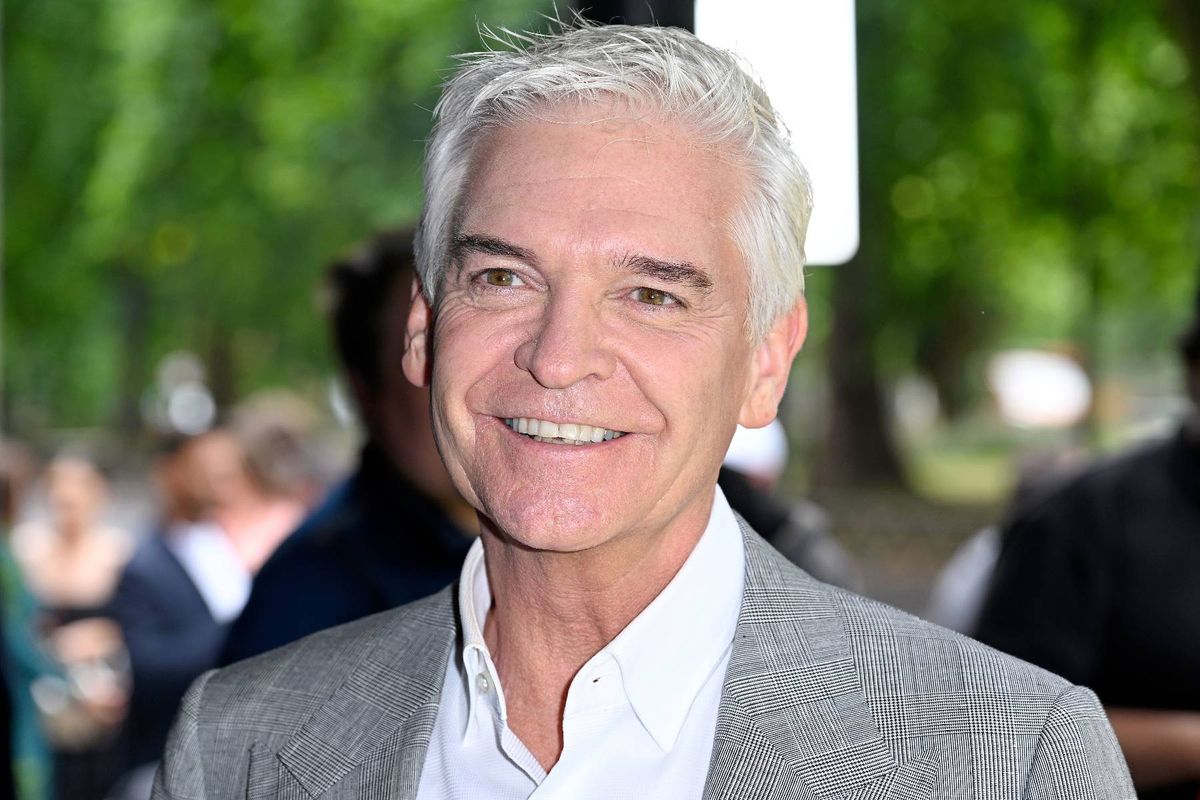 An individual with white hair, dressed in a light gray suit and a white shirt, is standing and smiling in an outdoor setting with a blurred background that includes several people and trees.