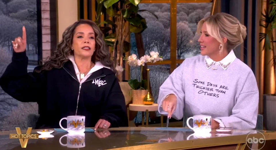 Two women sit at a talk show desk. One points upwards, mid-discussion; the other smiles. Both wear casual sweatshirts with text