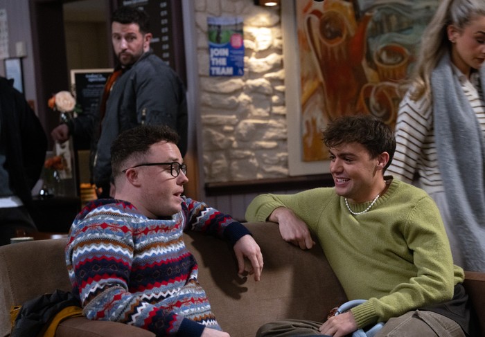 Ross and Kammy look over their shoulders towards Vinny and Ross, who are sat on a sofa in the cafe, in a scene from Emmerdale