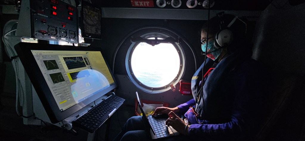 A woman in a blue facemask sits in an airplane, typing on a laptop with a large screen in front of her. The scene is lit by a round window in the middle of the frame.