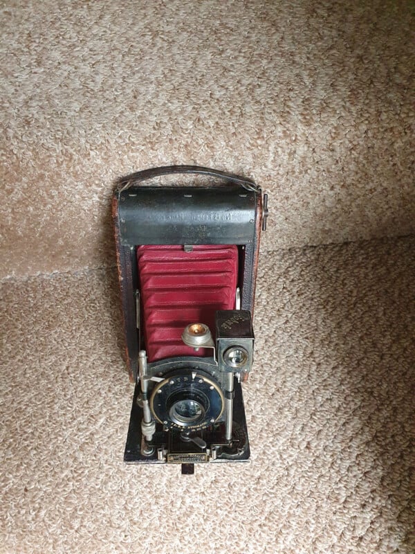An antique folding camera with a red bellows sits on a carpeted staircase, facing forward with its lens and viewfinder visible.