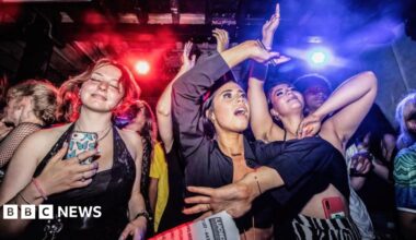 Three young women dancing in a Manchester nightclub with a crowd and flashing red and blue lights behind them