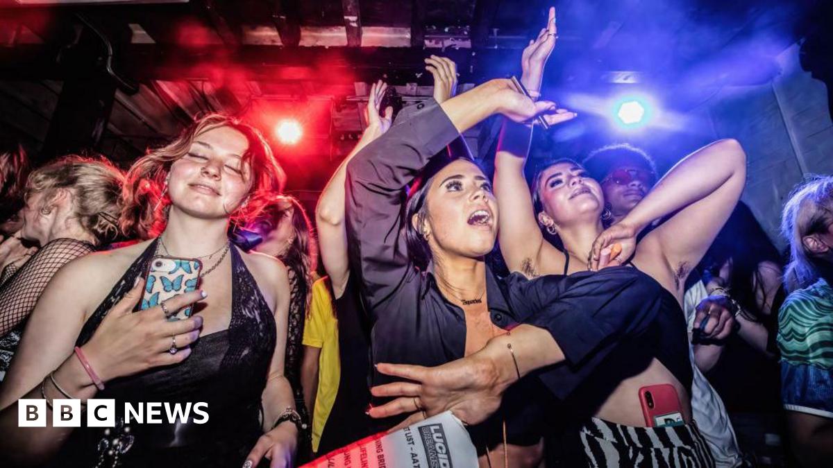Three young women dancing in a Manchester nightclub with a crowd and flashing red and blue lights behind them