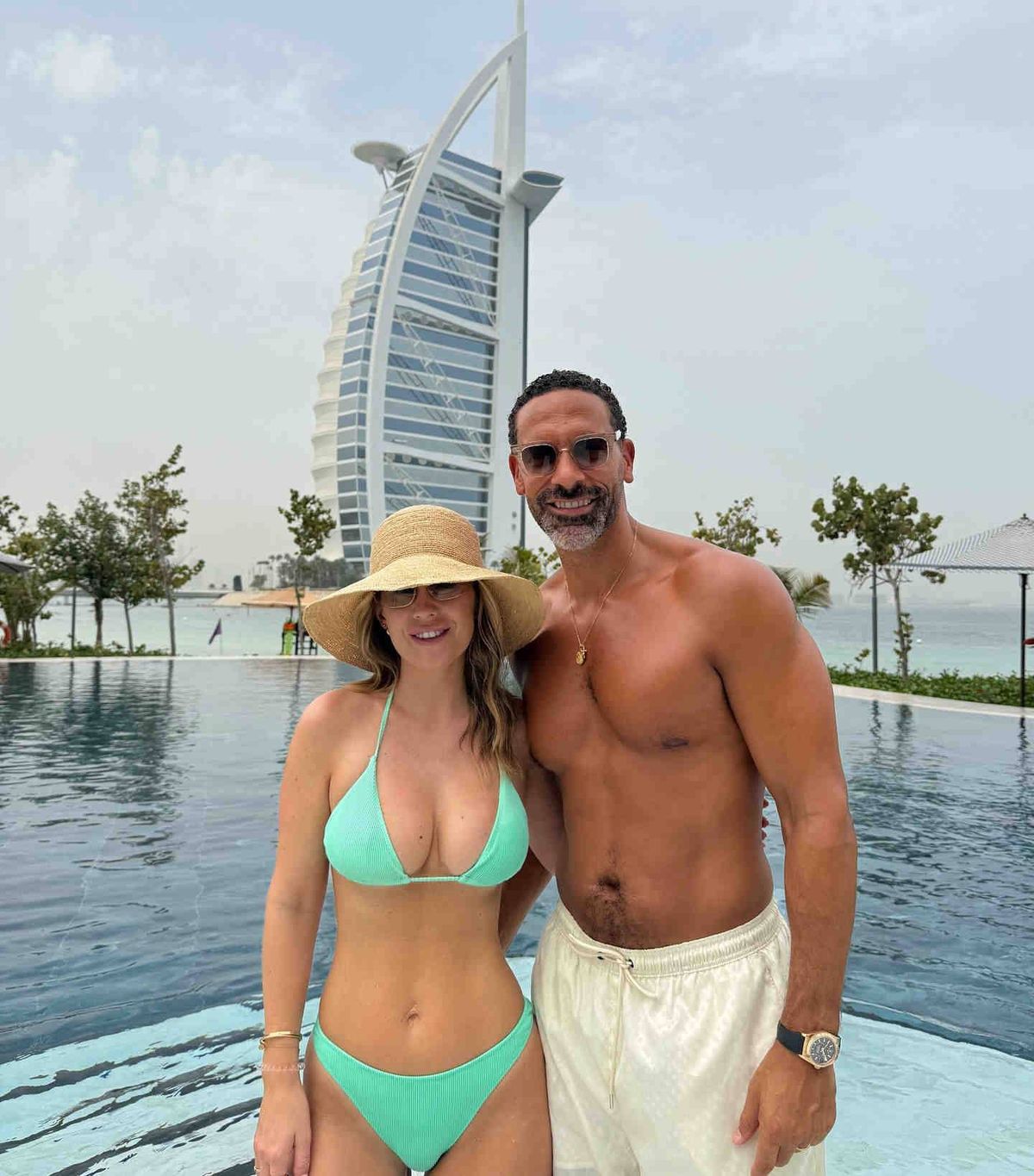 An adult male and female standing by a large swimming pool, possibly on vacation. The man is shirtless, while the woman is wearing a bikini and a hat. They are both smiling and appear to be posing for a photograph. Behind them are several trees and a high-rise building.