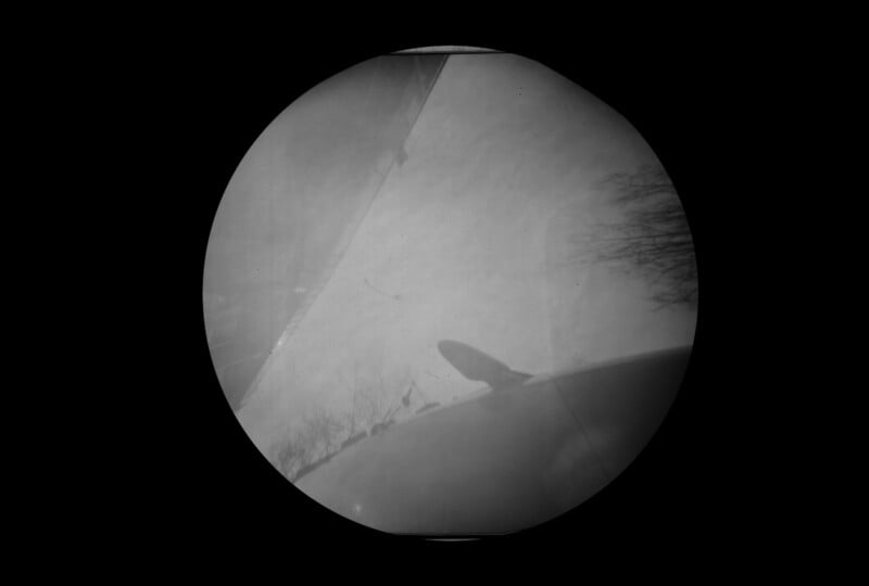 Black and white image viewed through a circular frame, showing the shadowy outline of a vehicle with an antenna, tree branches, and part of a wing or blade against a bright, cloudy sky.