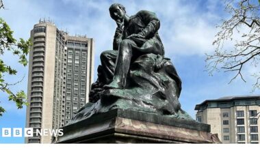 A bronze statue on a marble plinth, surrounded by the tall buildings on Park Lane. The statue depicts Lord Byron seated, with his head resting on his right hand in a pensive mood.