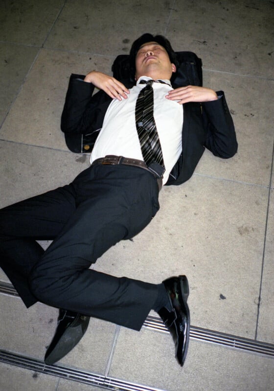 A man in a suit lies on his back on a tiled floor with his hands on his chest and eyes closed, using his backpack as a pillow.