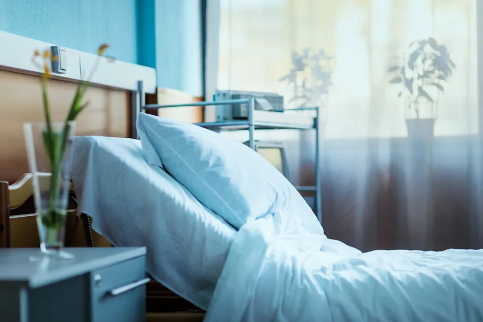 Hospital room with a neatly made bed, soft pillows, bedside table, and plants near a window