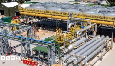 The United Downs power plant in Cornwall is shown, there are various silver pipes surrounding a central plant that has a series of large fan type structures surrounded by yellow pipes. There are trees in the background and the sky is blue.