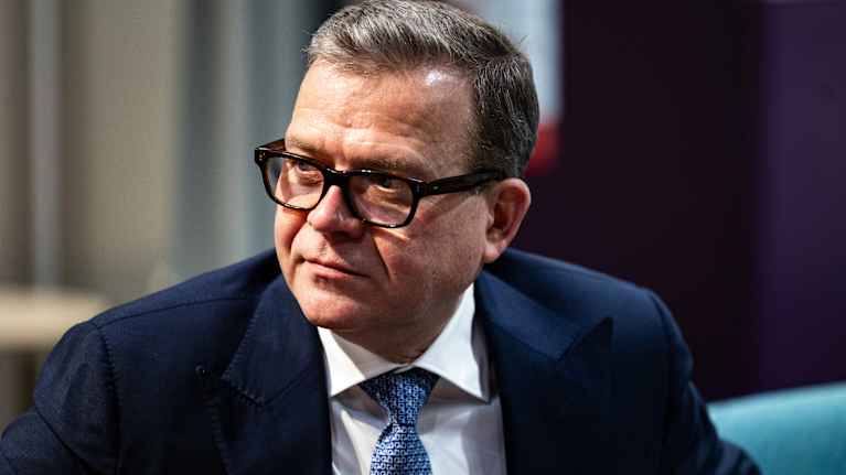 A man with short hair, wearing eyeglasses,, a dark blue suit jacket, white shirt and blue patterned necktie looking to the left.