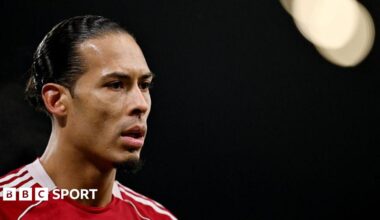 Close-up image of Virgil van Dijk in the left of the photograph. He has black hair, tied back, and is wearing Liverpool's red home shirt. The background of the image is black with a spotlight in the top right corner.