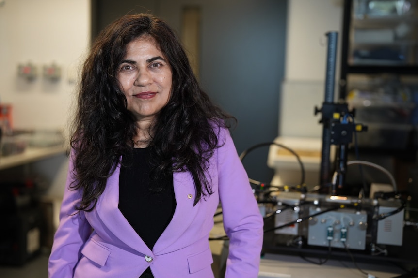 Woman with long black hair in purple jacket stand in a lab in front of objects.