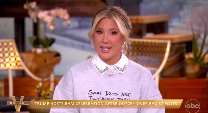 TV host sitting, wearing a sweater with text and a collared shirt, discusses a topic on a talk show. News headline displayed on the screen below