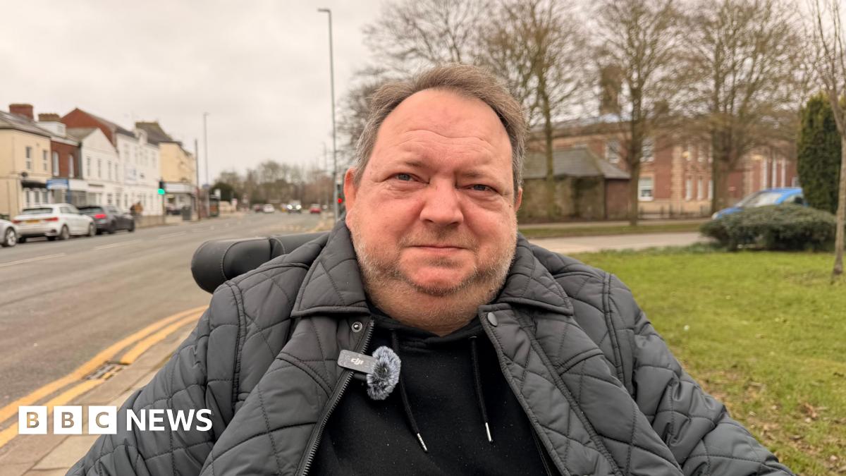A man sitting in a wheelchair on a path looking at the camera. He is wearing a black hoodie and a black padded jacket. He has short brown hair and stubble. Large buildings can be seen on either side of a road, with cars parked by the side of the road.