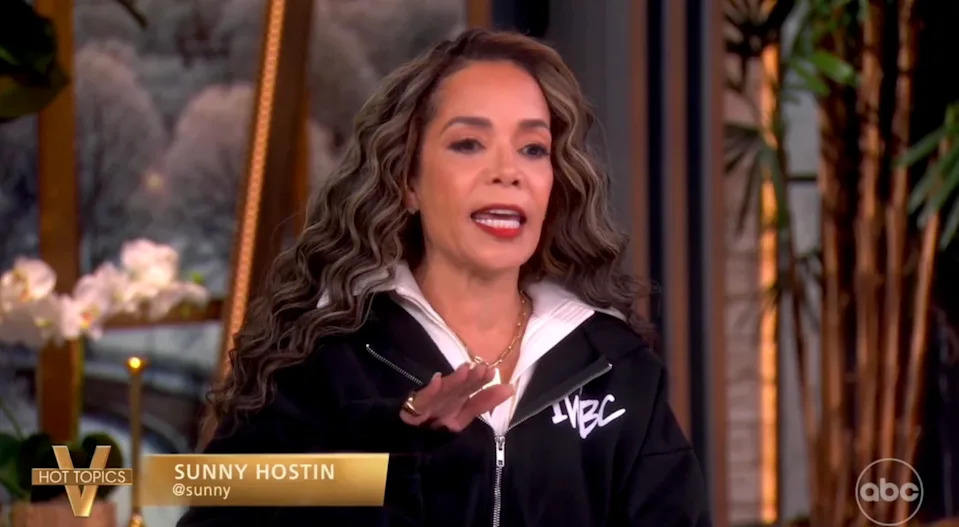 A person speaks on a talk show set, wearing a zippered jacket with "ABC" on it. Their hand is slightly raised. 