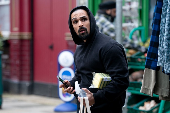 550930,TITLE:Eastenders Ravi, wearing a black hoody and carrying shopping, turns backwards in the EastEnders market.