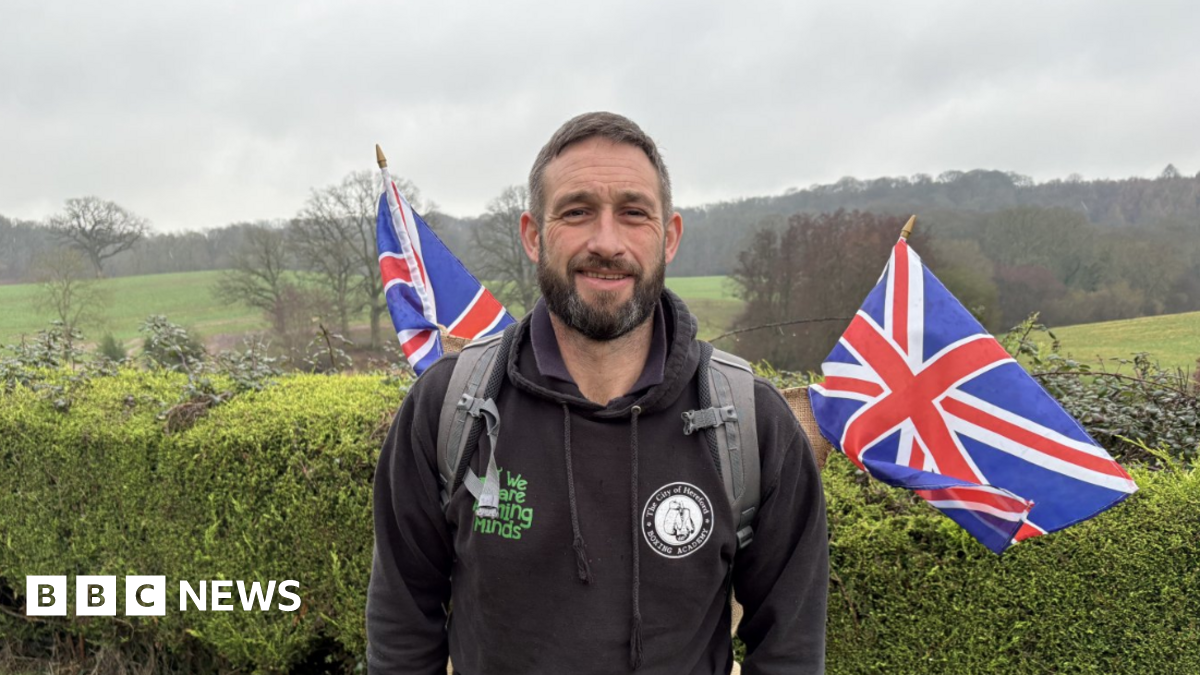 A man with dark hair and a dark beard wearing a black hoodie wears a grey rucksack on his back with two union jack flags coming from his back. He is stood in the countryside with a large hedge behind him.