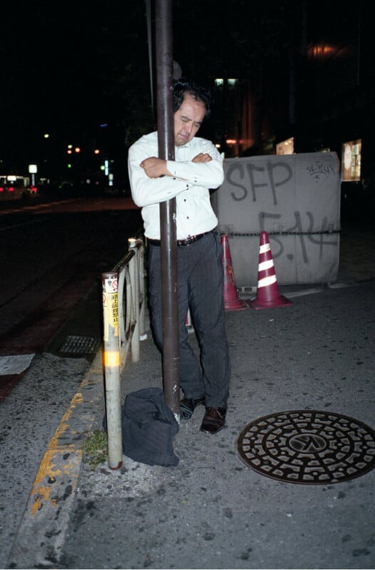 A man in formal clothes stands on a city sidewalk at night, leaning with his arms crossed against a pole. Nearby are traffic cones, a utility cover, a bag on the ground, and graffiti on a large container.