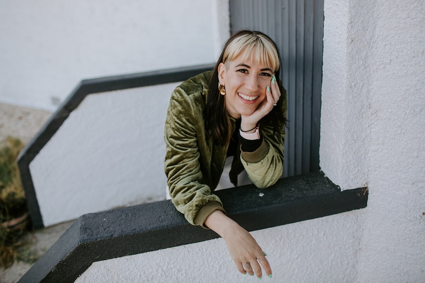 A woman with a bleach blonde fringe and black long hair leaning against handrail smiling for the camera