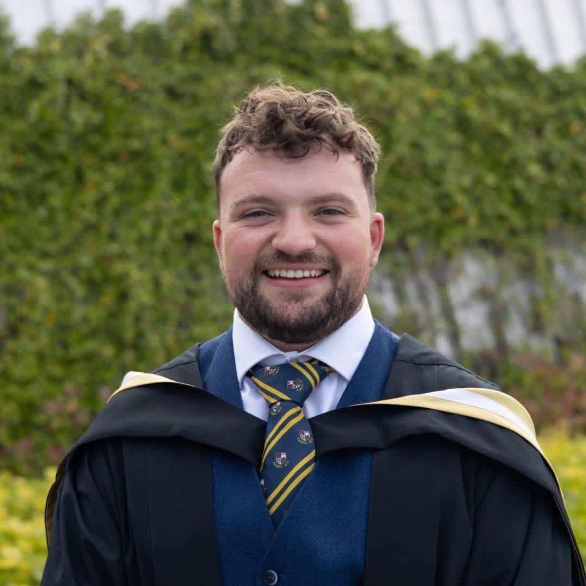An individual, likely a recent graduate, dressed in a graduation gown and tie, is standing with a joyful expression in front of a backdrop featuring foliage.