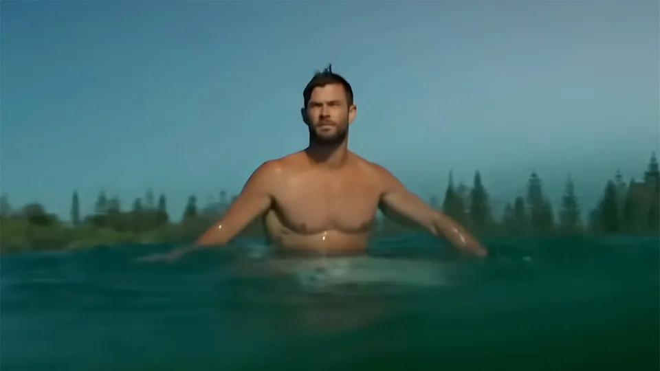 Chris Hemsworth screenshot of him surfing shirtless in bright blue water from video footage. 