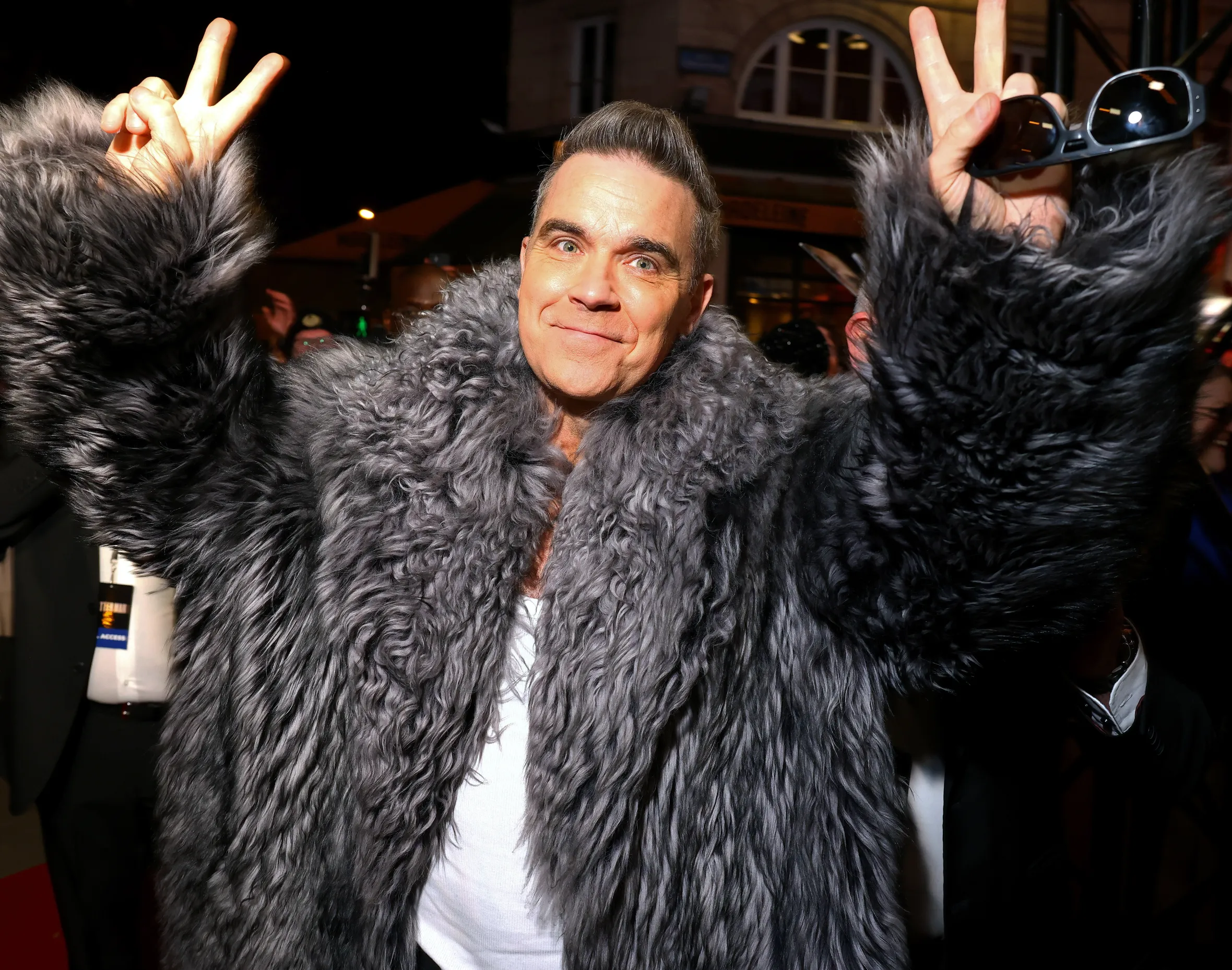 Robbie Williams wearing a gray fur coat and making peace signs with both hands at the "Better Man" special screening.