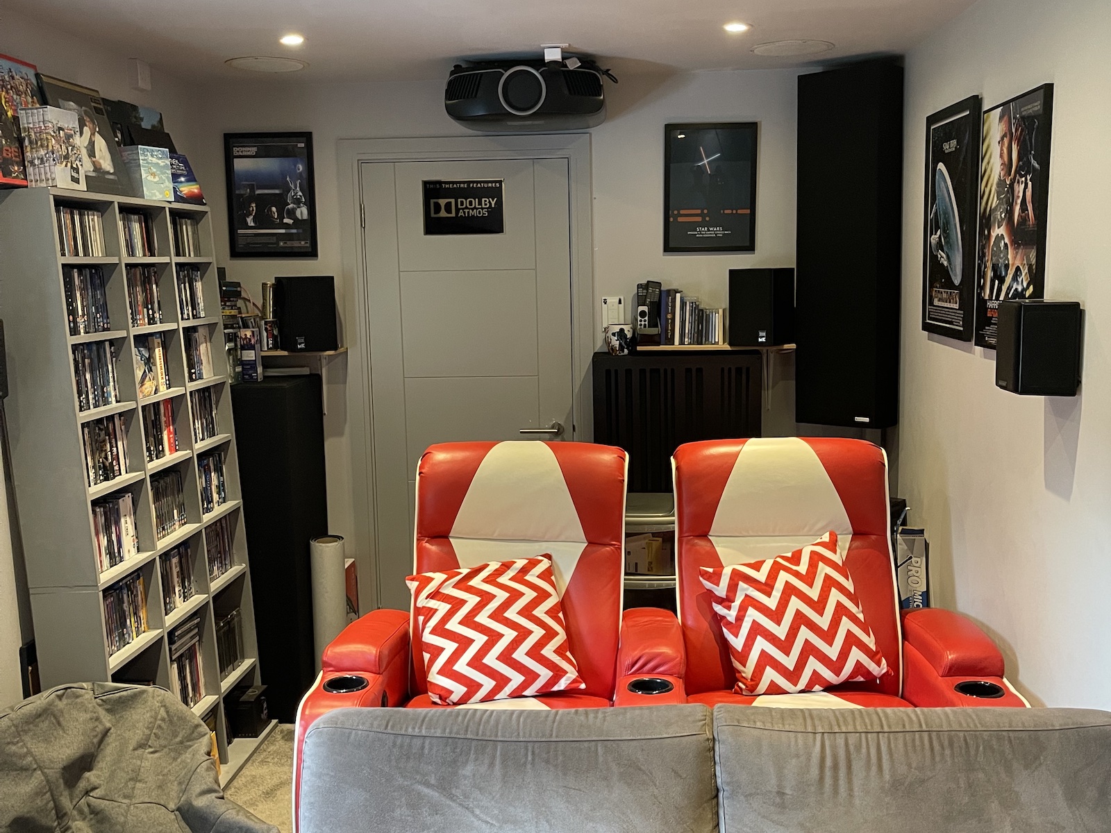 A photo of the back half of a dedicated home cinema room, showing the bright red seats, a ceiling-mounted projector, and shelves packed with movie discs