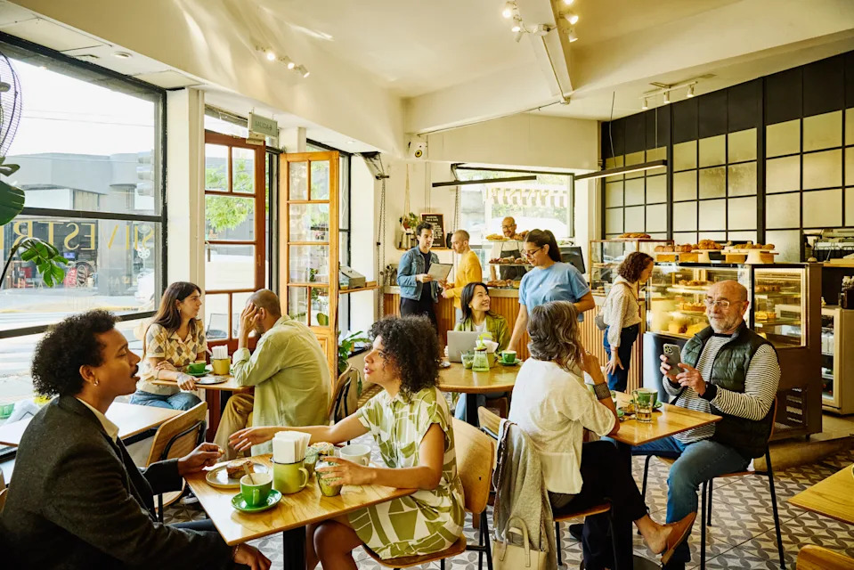 People sitting and chatting in a cozy café; others stand at a counter. The setting conveys a relaxed, social atmosphere typical of work breaks