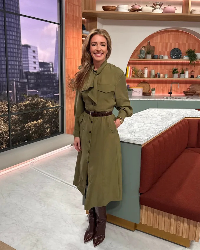 a person wearing a long olive green dress with a tied collar stands in a modern kitchen setting