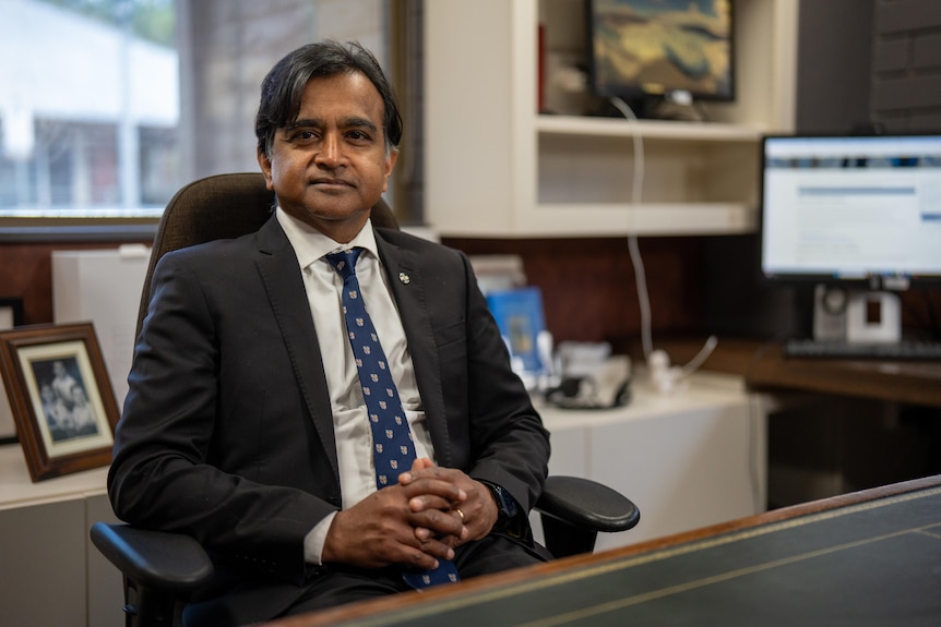 A man in a business suit sits in an office chair behind a desk. A lit up computer screen next to photos and shelves in the back