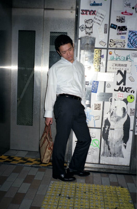 A man in business attire stands slouched with his eyes closed, holding a brown bag, in front of a wall covered with various stickers and graffiti near a subway or building entrance.