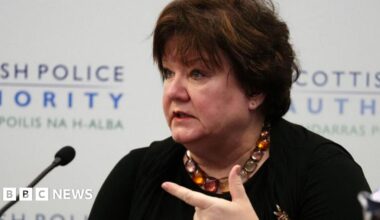 Fiona McQueen, who has short black hair, speaks into a microphone. She is wearing a black top and with her left index finger pointing to her right.