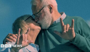 Two young baristas, wearing black aprons with the Dialogue cafe logo on the front - which includes text in white and a large purple heart - stand in front of the café's windows.