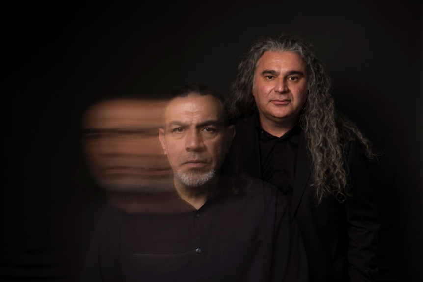 Khaled Sabsabi, early 60s, Lebanese Australian, frowns, as Michael Dagostino, with long grey hair, stands behind him.