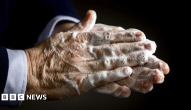 A close up image of the hands of a person who is wearing a suit. The picture has been taken while they are washing their hands and there is soap residue which is yet to be washed off.