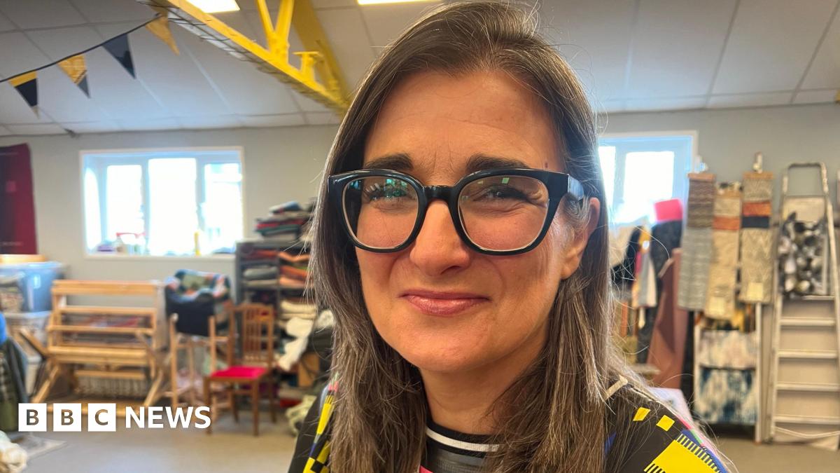 Melissa Santiago-Val is sat in an open spaced room. She has long brown hair and is wearing black frames glasses and a patterned top. Behind her are art supplies and a silver ladder.