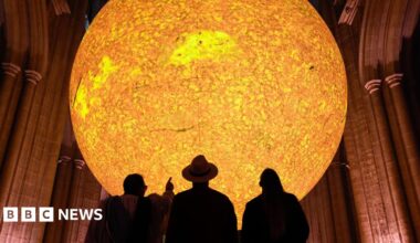 A large sun sculpture with a bright yellow and orange glow is suspended above a church-like building. The silhouettes of three people can be seen looking up at it, with one person pointing at it.