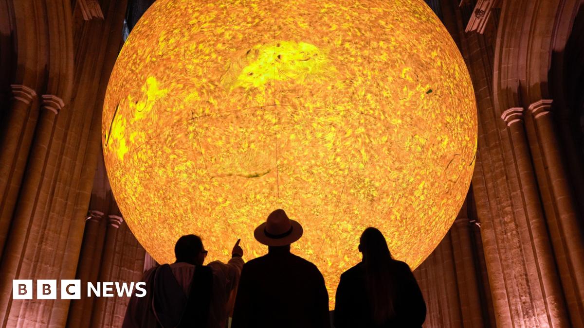 A large sun sculpture with a bright yellow and orange glow is suspended above a church-like building. The silhouettes of three people can be seen looking up at it, with one person pointing at it.