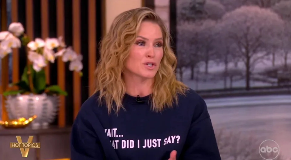 Person speaking on a TV show set, wearing a sweatshirt with the text: "Wait... What Did I Just Say?"