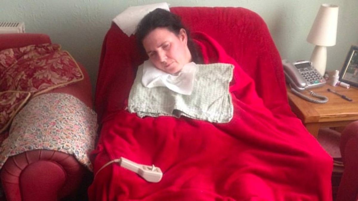 An individual is resting on a chair with a red cushion and blanket, accompanied by a white pillow and a device that appears to be a remote control. A lamp is situated on the right side, and a table with a telephone is nearby.