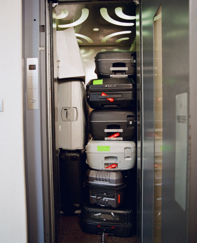 A small elevator packed from floor to ceiling with various stacked black and gray suitcases, leaving no room for passengers. The elevator doors are partially open.