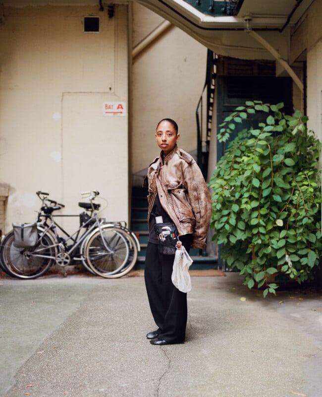A person stands outdoors near a leafy bush and two parked bicycles, wearing a brown leather jacket, black pants, and holding a white bag. A beige wall and metal staircase are in the background.
