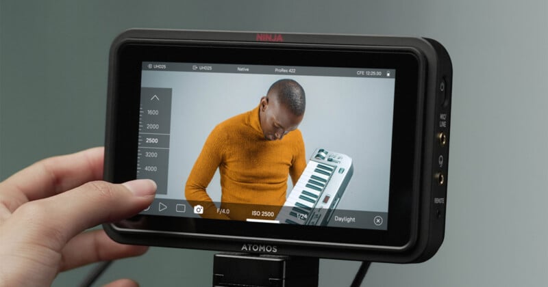 Close-up of a hand operating an Atomos Ninja camera monitor displaying a video of a person in an orange sweater holding an electronic keyboard.